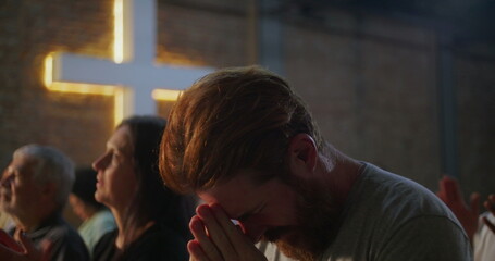 Man bowing head in prayer, hands clasped, deeply engaged in spiritual practice in a church, illuminated cross on a brick wall in the background