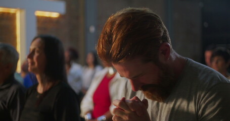Man bowing head in prayer, hands clasped, deeply engaged in spiritual practice in a church, illuminated cross on a brick wall in the background