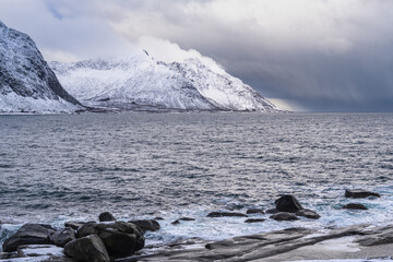 rocks on shore of ocean, stony sand gravel beach with waves, fjord landscape with high snowy...