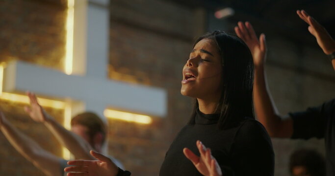Focused woman with raised hands in prayer, deep spiritual connection, Christian faith, worship gathering, illuminated cross in background, unity in worship, religious devotion
