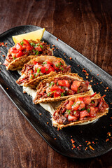 Four crispy tacos with shredded meat, diced tomatoes, onions, and cilantro on a black plate with a lime wedge. Garnished with chili flakes, served on a wooden table