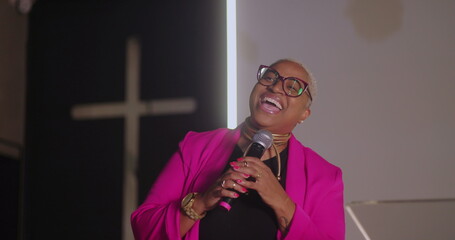 Smiling woman in bright pink blazer speaking passionately on stage, holding microphone, Christian...