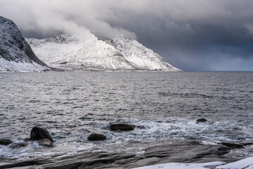rocks on shore of ocean, stony sand gravel beach with waves, fjord landscape with high snowy...