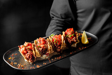 A person in a black shirt holds a black plate with four tacos filled with shredded meat, diced tomatoes, and herbs. A lemon wedge is placed on the side. The tacos are garnished with spices