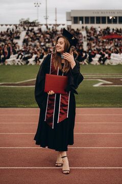A young woman stands in a stadium wearing a gown with a diploma. One hand touches her hair, the other holds a red folder. The emotion is calm, a moment of relaxation.