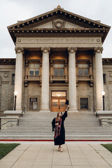 Obraz premium Graduation photo from the University of Redlands, a girl in a robe holds her graduation hat with her hand raised to the mountain, standing in front of the university entrance.