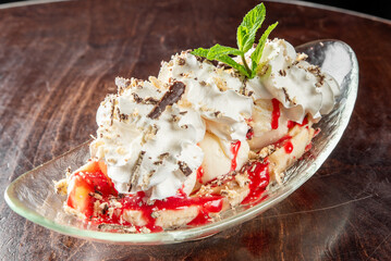A delicious banana split dessert with whipped cream, chocolate shavings, and fresh mint, drizzled with strawberry sauce. Served in a glass dish on a wooden table, perfect for food photography