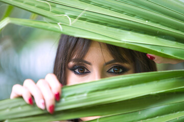 Pretty young brunette woman clutches a palm leaf in her hands and looks through it. The woman is...