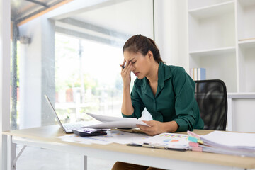 A woman is sitting at a desk with a laptop and a calculator. She is writing on a piece of paper and...