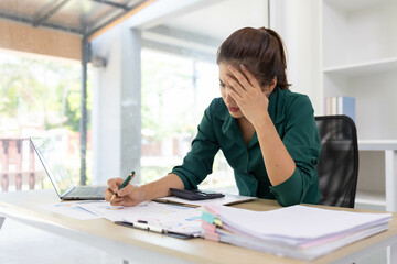A woman is sitting at a desk with a laptop and a calculator. She is writing on a piece of paper and...