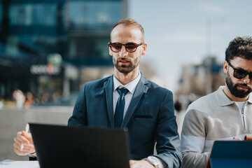 Professionally dressed individuals using laptops outdoors in an urban area, showcasing modern work culture and collaboration.