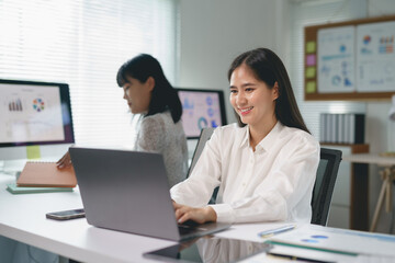 Obraz premium Young Asian businesswoman smiling and working on laptop in modern office with charts and graphs displayed on computer screen
