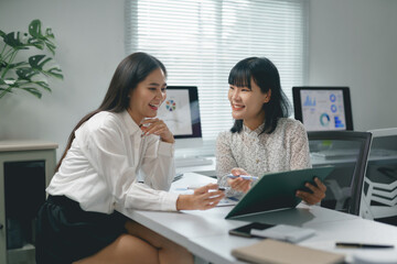 Two asian businesswomen are discussing marketing strategy while working together and smiling in modern office