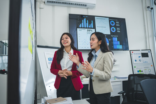Two businesswomen discussing and analyzing data displayed on a monitor and whiteboard during a presentation in a modern office