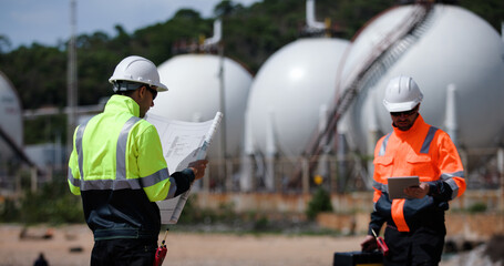 Two man industrial engineer with green and orange safety suit working with blueprint drawing at Refinery and gas petrochemical plant