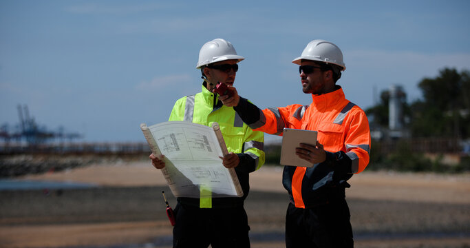 Two male power plant engineers work with a blueprint and a digital tablet by the seaside at a refinery and gas petrochemical plant