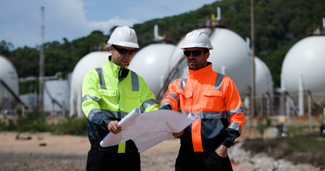 Two man industrial engineer with green and orange safety suit working with blueprint drawing at Refinery and gas petrochemical plant