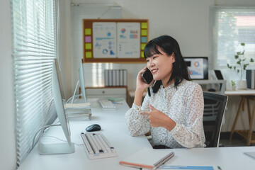 Young Asian businesswoman engaging in a phone conversation while pointing at her computer screen, focused on tasks in a modern office setting