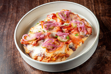 A delicious open-faced sandwich with melted cheese, crispy bacon, pickles, and pickled red onions, garnished with fresh parsley, served on a white dish atop a rustic wooden table.
