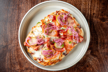 A delicious open-faced sandwich with melted cheese, crispy bacon, pickles, and pickled red onions, garnished with fresh parsley, served on a white dish atop a rustic wooden table.
