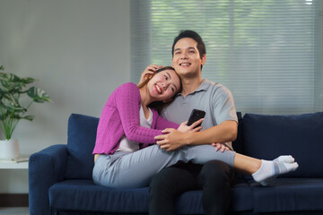 Obraz premium Happy asian couple is spending time together on the sofa at home, using a smartphone and smiling