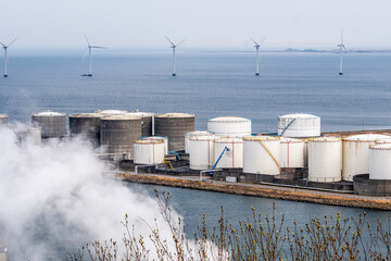 Coastal fuel storage site with large tanks and drifting smoke, emphasizing fossil energy infrastructure and industrial landscape near the sea.