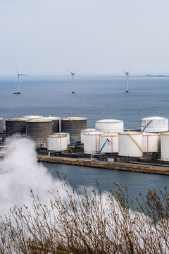 White steam and smoke rising from storage tanks by the coast, with wind turbines visible in the background for energy and environmental contrast themes.