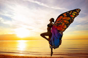 Ballerina jumping with Colorful Butterfly Wings on the Beach in Sunset Sunshine. Ballet Dancer Side View Silhouette at Sea. Magical Transformation. Woman dancing at Seashore. Rebirth and Meditation