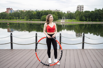 A girl in a red tank top and sweatpants does exercises and acrobatic tricks with a hula hoop on a...