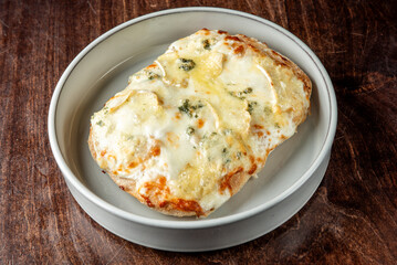 Golden, baked bread topped with melted cheese and aromatic herbs, served on a white plate over a rustic wooden table. The warm, crispy texture and gooey cheese