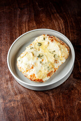Golden, baked bread topped with melted cheese and aromatic herbs, served on a white plate over a rustic wooden table. The warm, crispy texture and gooey cheese