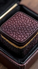 Fototapeta premium A small, textured, burgundy jewelry box with a gold accent, resting inside an open case on a wooden surface.