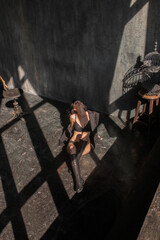 young brunette girl in black lingerie, jacket and boots is sitting fashion in sun lights on the floor and looking up at dark wall background in art workshop interior. lifestyle concept, free space