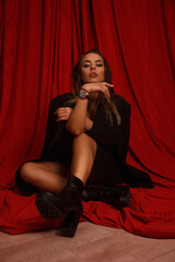 young blonde girl in black jacket and boots is sitting fashion with hand near face and looking straight with flirt on the red fabric background. fashion concept, free space