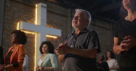 Elderly man clapping during church worship, illuminated cross in background, diverse group...
