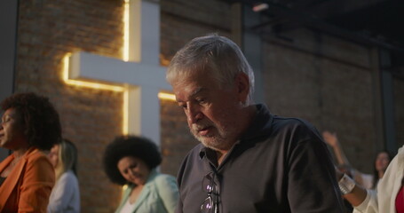 Close-up of elderly man looking up during prayer in church, illuminated cross in background, expression of deep spirituality, faith, and religious connection
