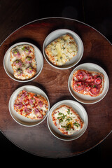 Top view of five assorted mini pizzas served on white plates, arranged in a circular pattern on rustic wooden table. Each pizza features different toppings, including cheese, pepperoni, and vegetables