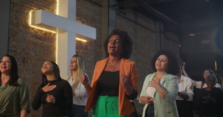 Women singing and worshiping together in diverse church community, standing near illuminated cross,...