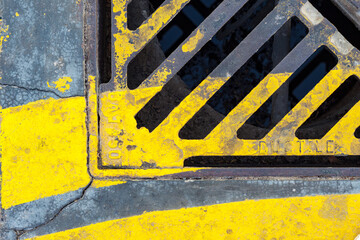 A weathered yellow and gray drainage grate, showcasing the texture and rust of urban infrastructure.