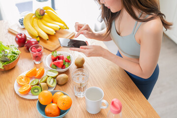 Asian beauty woman in stylish sportswear prepares a healthy fruit drink in her modern home kitchen while smiling and taking a photo with her smartphone to share on social media, embracing wellness