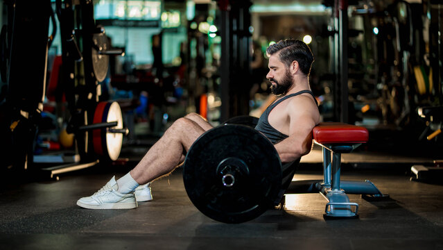 Strong man in black tank top preparing for hip thrust exercise with barbell on bench in gym, focused on glute and lower body training with proper form