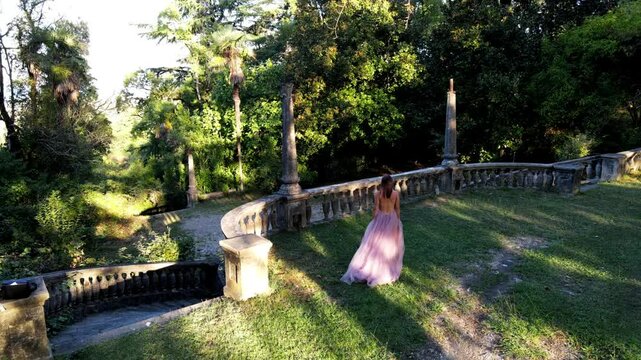 Romantic young European woman in blush pink open-back maxi dress walks through enchanted green forest and steps onto an old stone balcony staircase. Dreamy cinematic slow motion scene in nature.