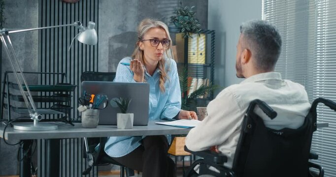 A focused female employer explains and questions seriously, talking to a man in a wheelchair during job interview in a modern office.