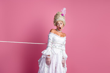 Graceful aristocratic lady tied with rope in baroque costume expressing humorous surprise on pink background