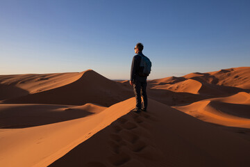 brave and strong man traveler with backpack standing in Sahara desert sand dunes at sunset, travel adventure outdoor