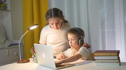 Boy in headphones writes on paper while girl helps studying. Girl explains student tasks at laptop. Child learning in home interior. Writing exercise continues kid and girl. Headphones improve focus. - Powered by Adobe