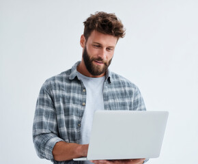 A bearded man in a plaid shirt uses his laptop, deep in thought, indoors.
