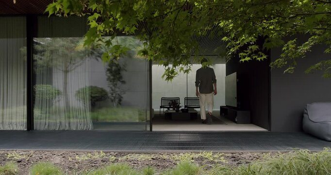 A stylish man dressed in smart casual attire walks towards a modern house surrounded by lush greenery and elegant transparent curtains. Modern Home Exterior with Man Walking into Stylish House
