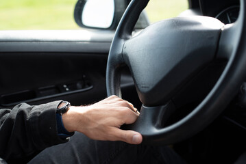 Close-up of a man's hands gripping the steering wheel while driving a car. Green foliage is visible through the window, suggesting movement and a natural outdoor environment. 
