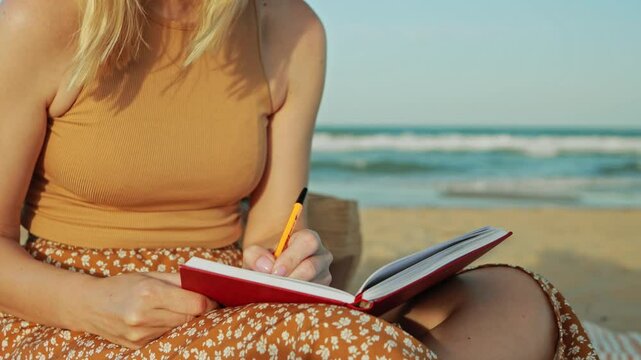 Blond woman writing in red notebook, wearing light summer dress, seated on sandy shore, adjusting straw hat, capturing peaceful coastal moment with yellow pen.Summer concept